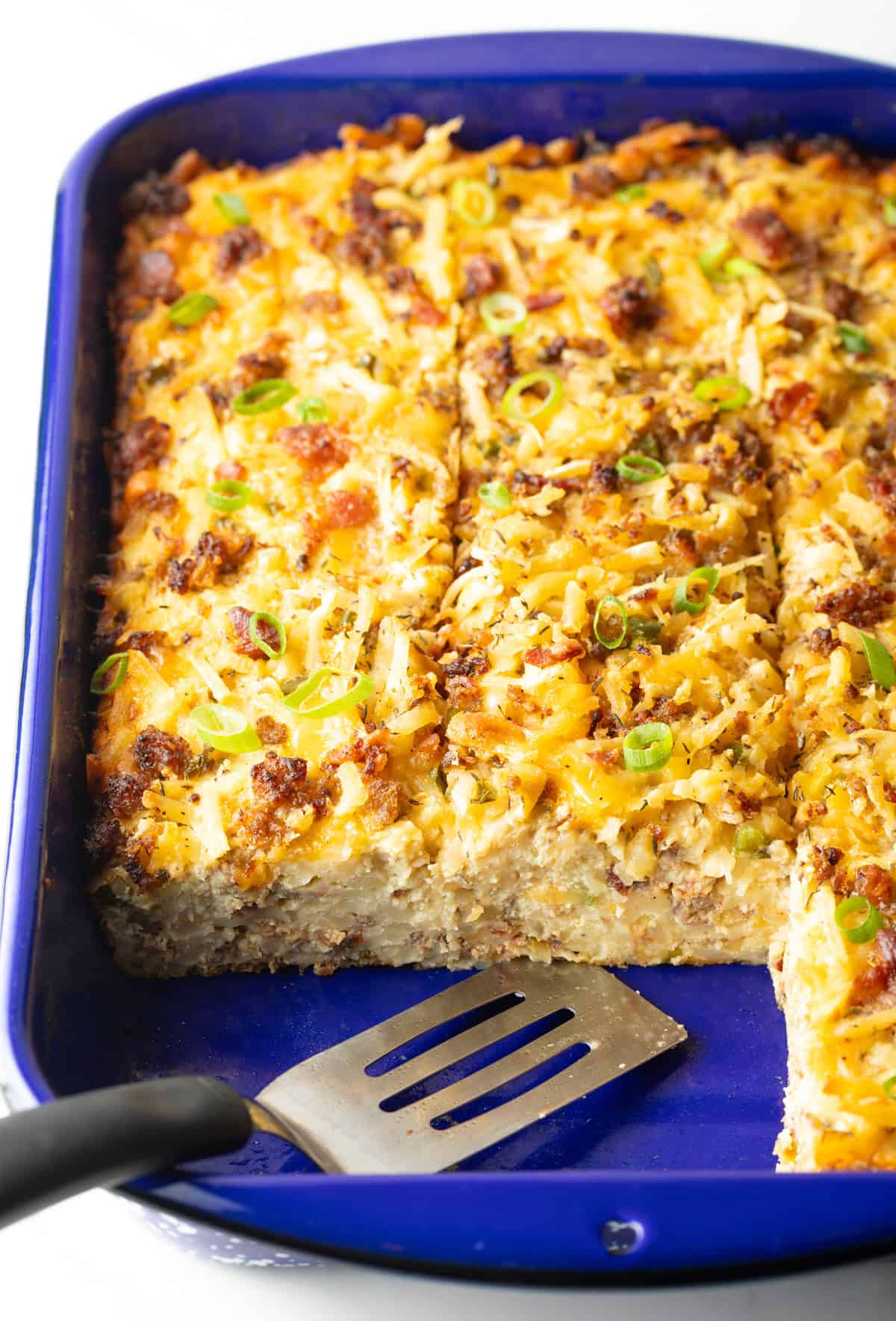 Sliced breakfast hashbrown casserole in a blue baking dish with two pieces missing, with a metal spatula.