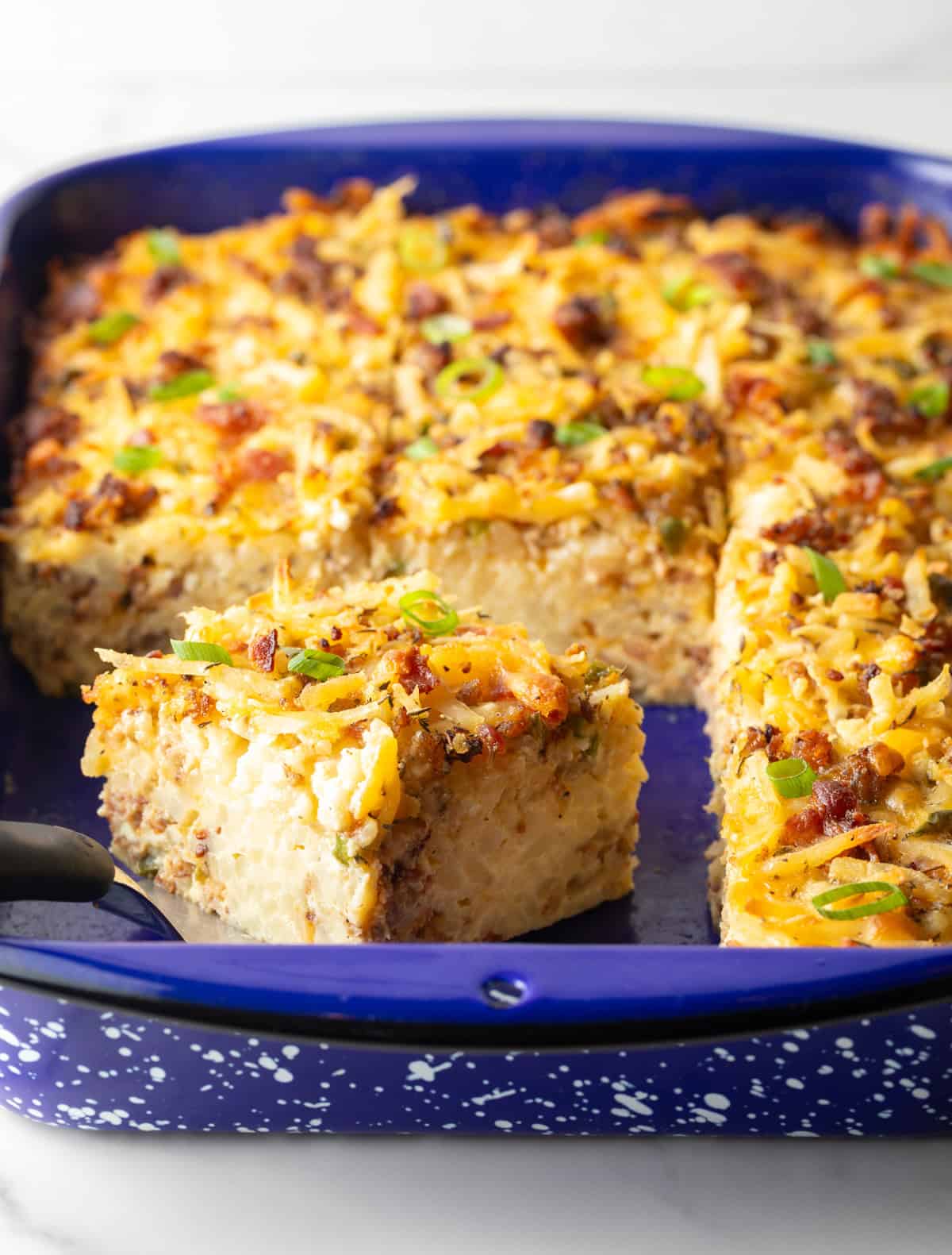 A hash brown breakfast casserole in a blue baking dish, wish a slice being held by a metal spatula.