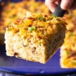 A slice of hashbrown breakfast casserole is being held by a metal spatula above a blue baking dish.