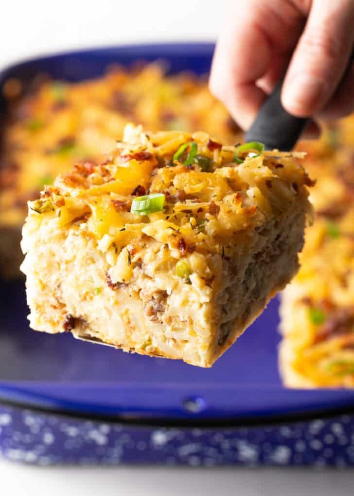 A slice of hashbrown breakfast casserole is being held by a metal spatula above a blue baking dish.