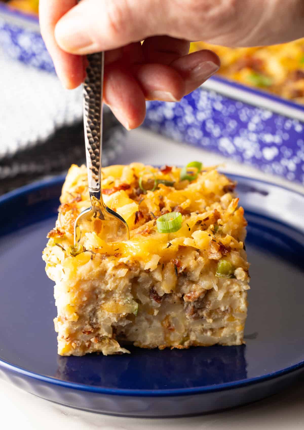 A slice of hashbrown breakfast casserole on a blue plate, with a fork taking a bite out of it.