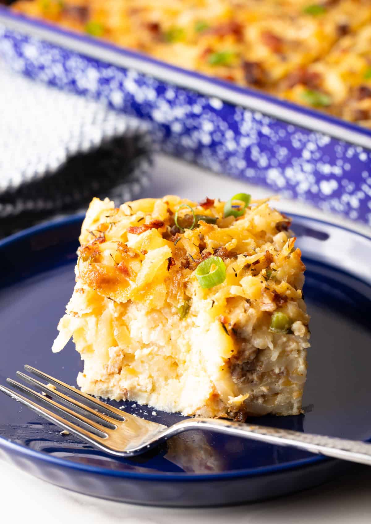 A slice of breakfast casserole with hash browns on a blue plate, with a fork and a bite taken out of it.