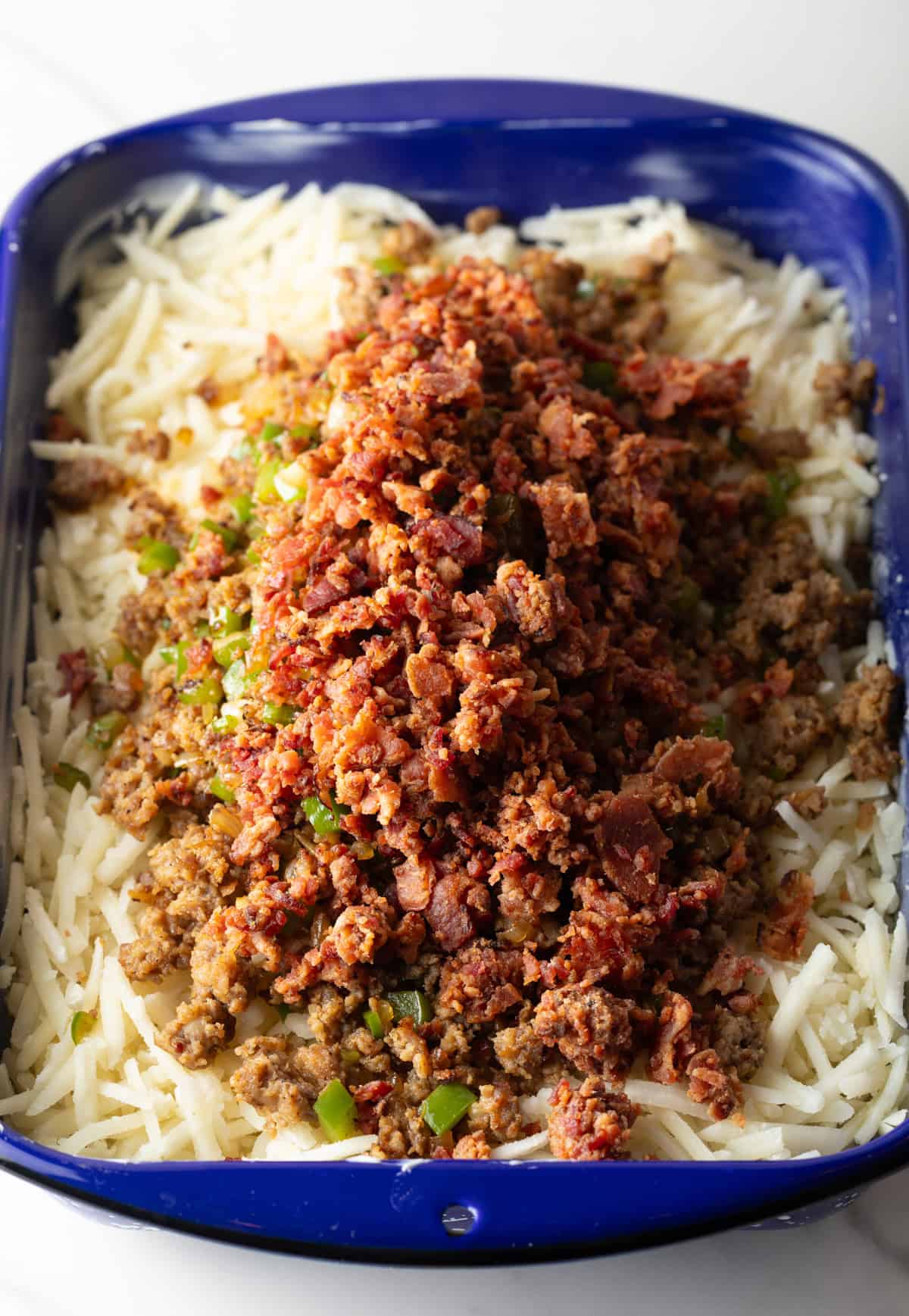 Frozen hash browns are underneath cooked sausage and bacon in a blue casserole dish.