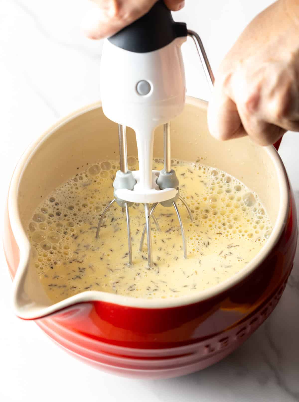 A whisk is mixing an egg and cream mixture in a red bowl.