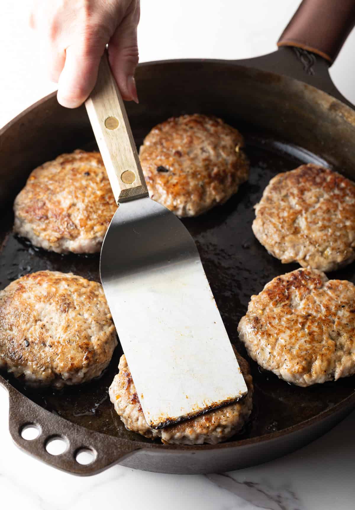 Homemade breakfast sausage patties in a black skillet, with a metal spatula pressing one of the patties.