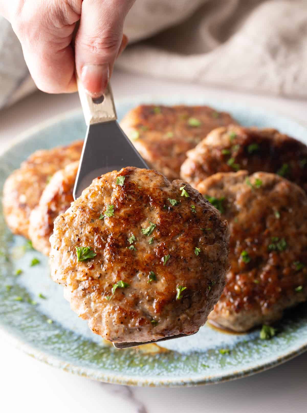 A metal spatula holding a breakfast sausage patty over a plate of homemade breakfast sausage.