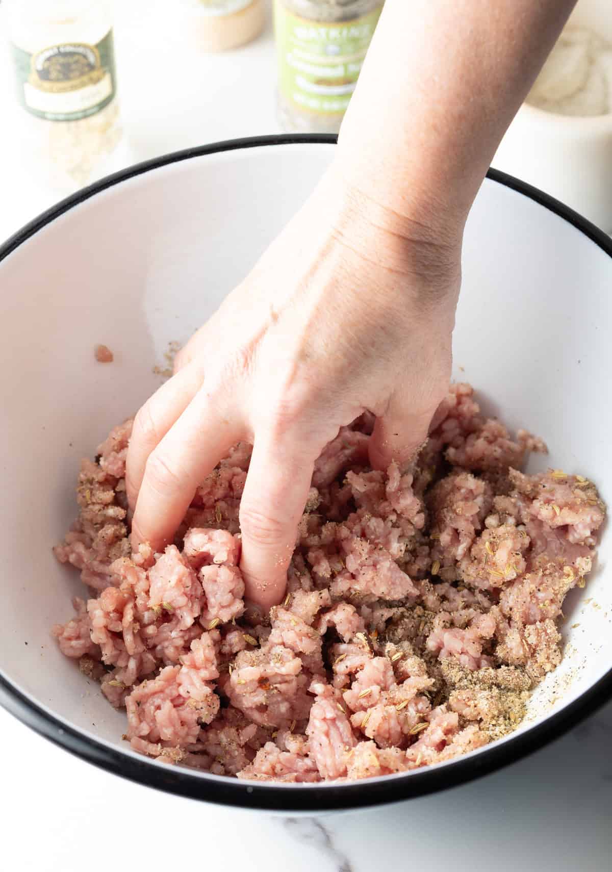 A hand is mixing a ground pork and seasoning mixture together in a white bowl.