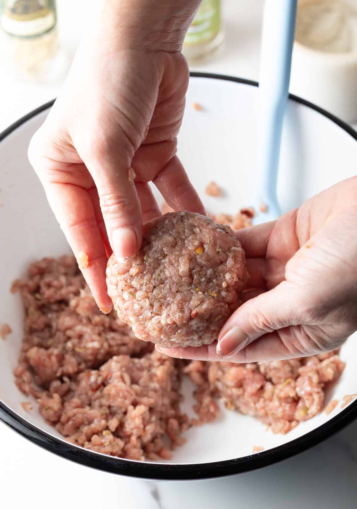 A hand holding a raw breakfast sausage patty over a white bowl filled with the homemade breakfast sausage mixture.