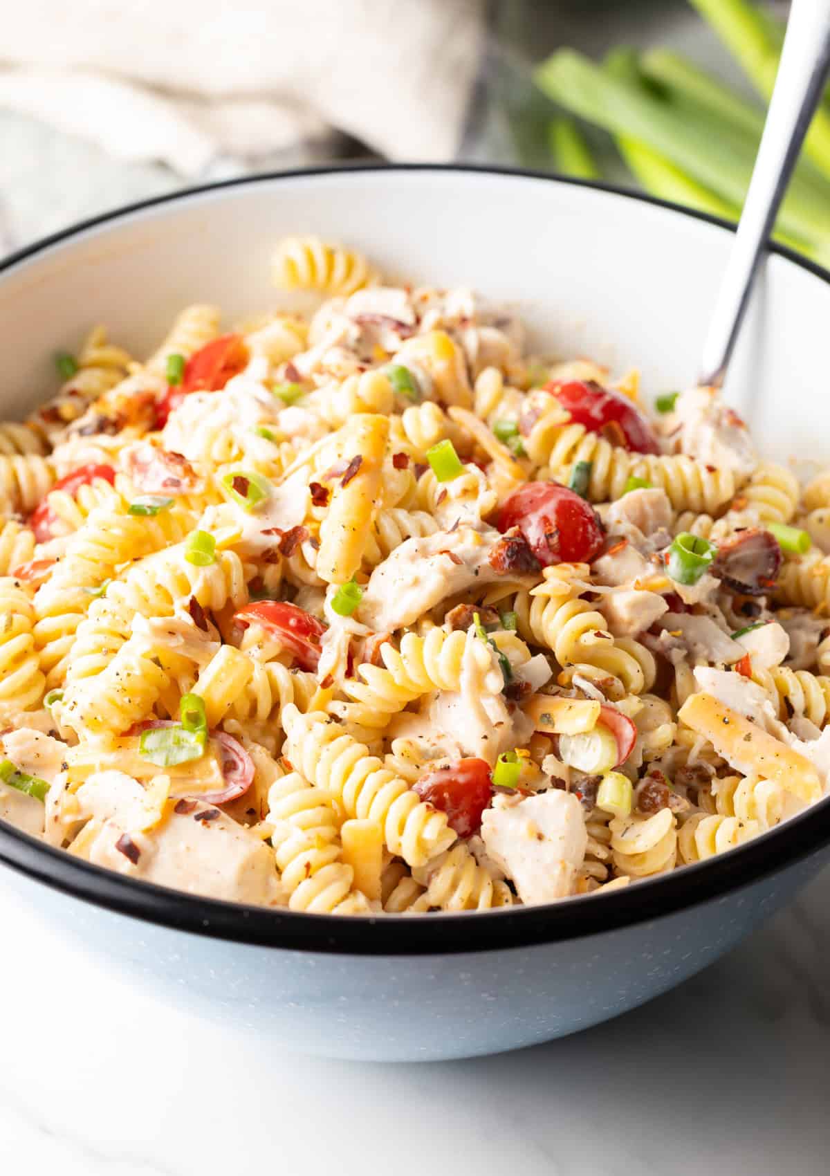 A metal spoon is stirring chicken bacon ranch pasta salad in a white bowl.