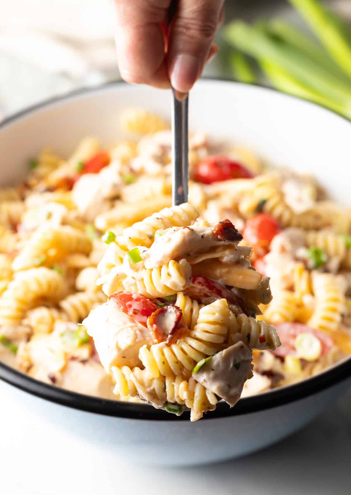 A metal spoon is holding a bite of chicken bacon ranch pasta salad above a bowl filled with the pasta salad.