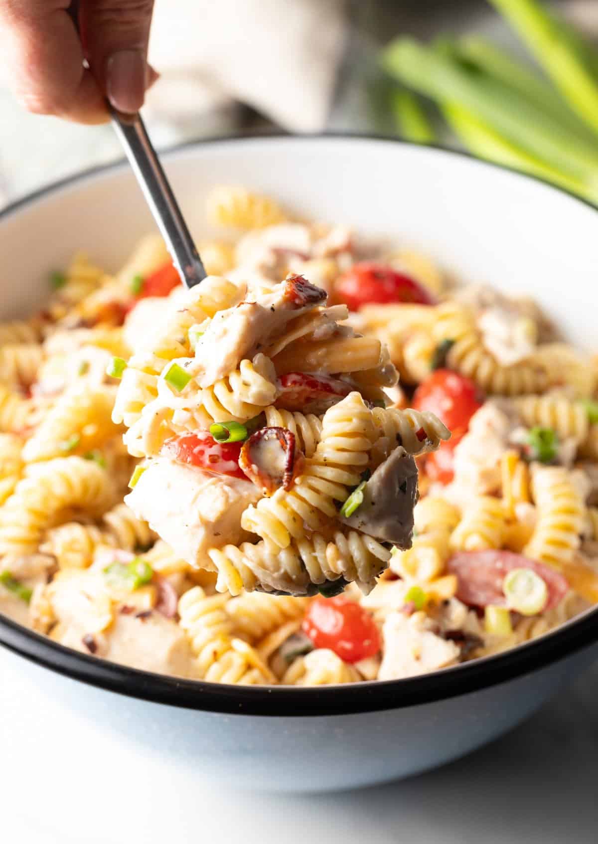 A metal spoon is holding a scoop of chicken bacon ranch pasta salad above a bowl filled with the pasta salad.