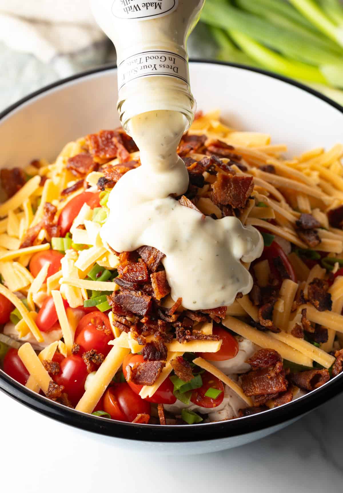 Ranch dressing is being poured over a bacon topped chicken bacon ranch pasta salad, ready to be mixed.