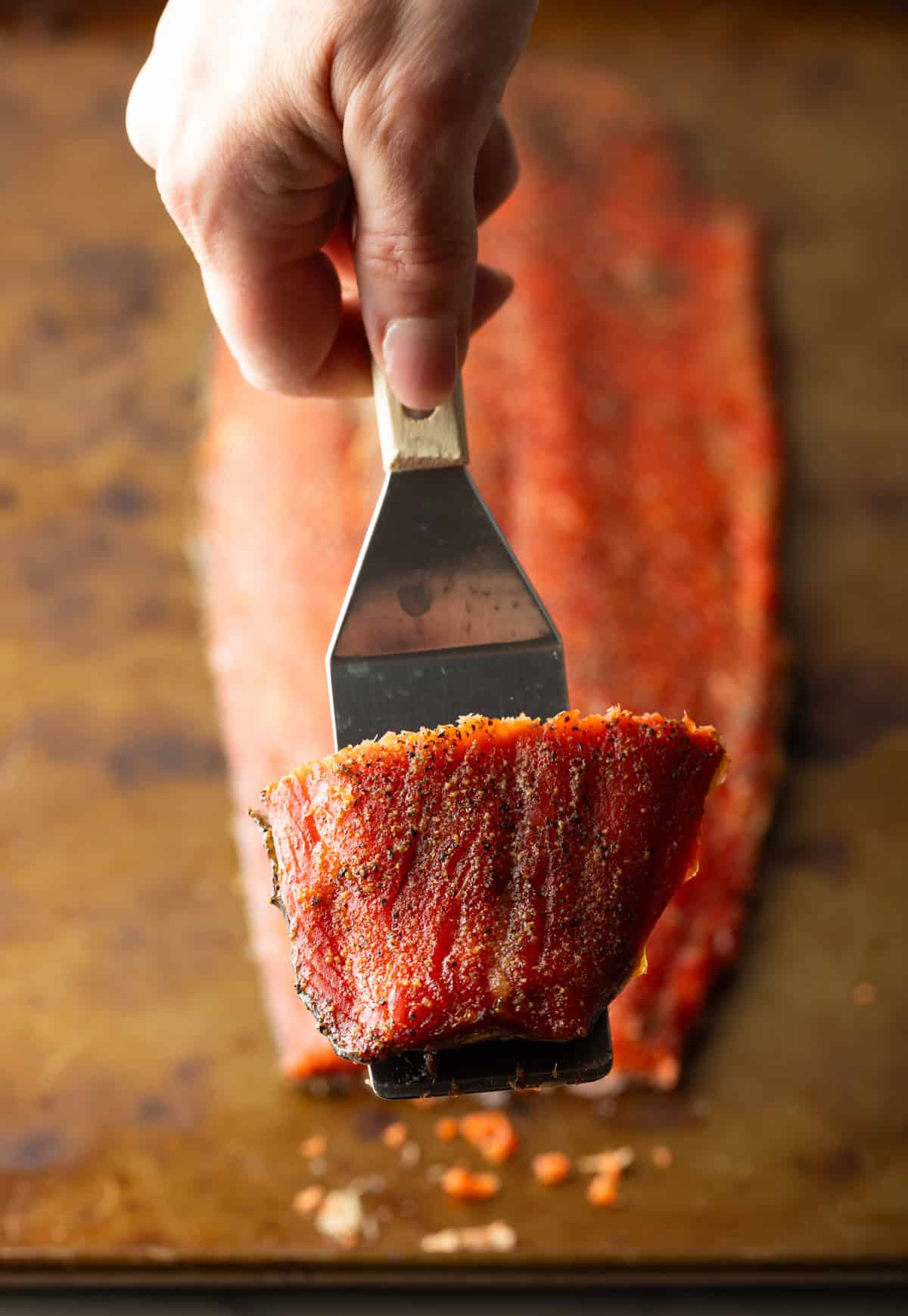 A metal spatula is holding a slice of easy cold smoked salmon above the full fillet of salmon.