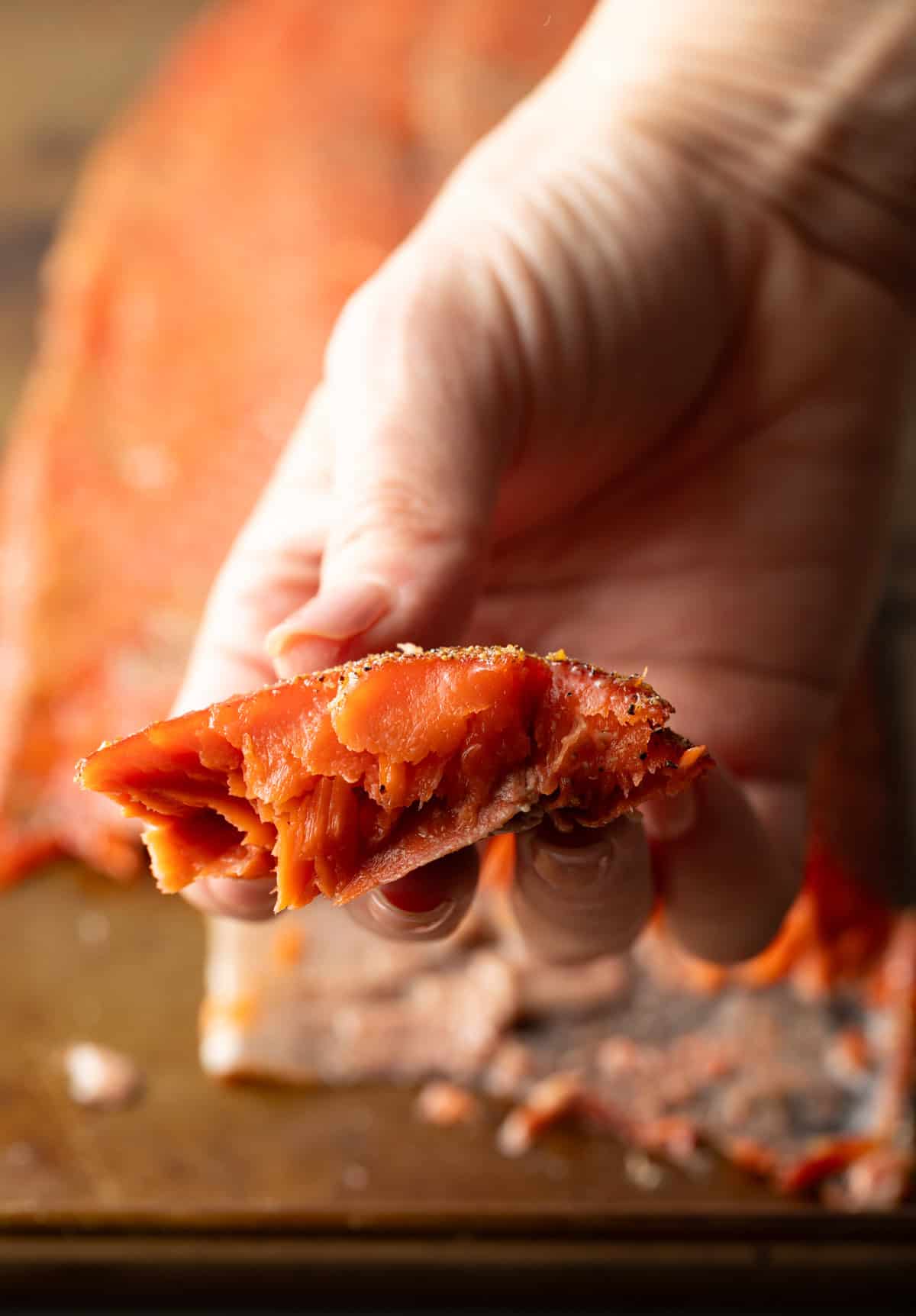 A hand holding a piece of smoked salmon, above the full fillet of cold smoked salmon.