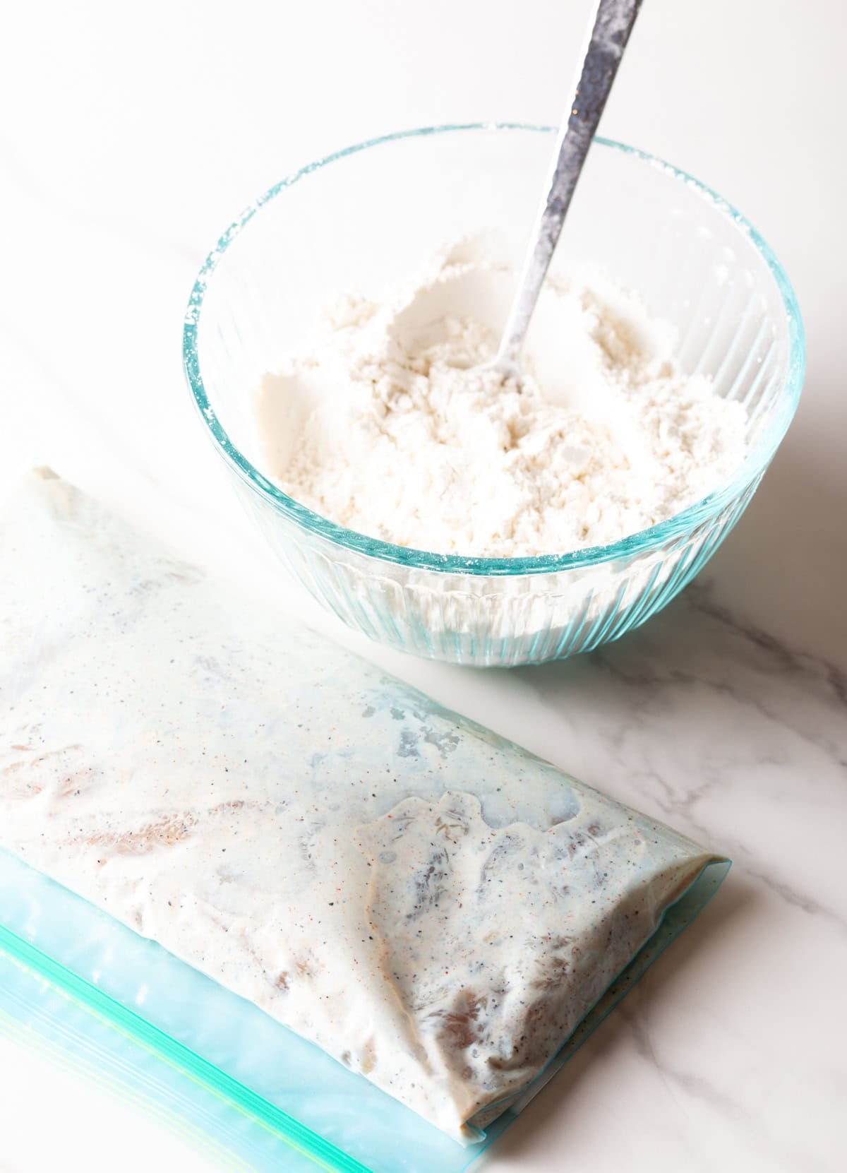 A glass bowl filled with a flour mixture sitting next to a bag of raw chicken tenders marinating in a buttermilk mixture.
