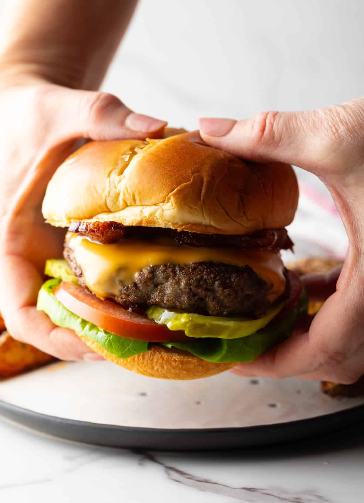 A hand is holding a fully assembled deer meat burger topped with cheese, lettuce, tomato, and pickles.