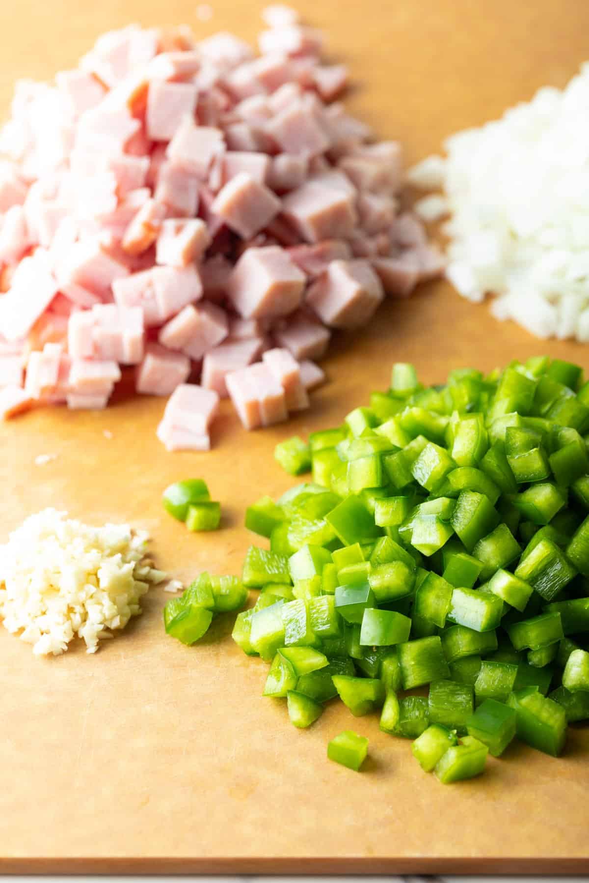 Dice ham, green bell peppers, and garlic sitting in piles on a wooden cutting board.