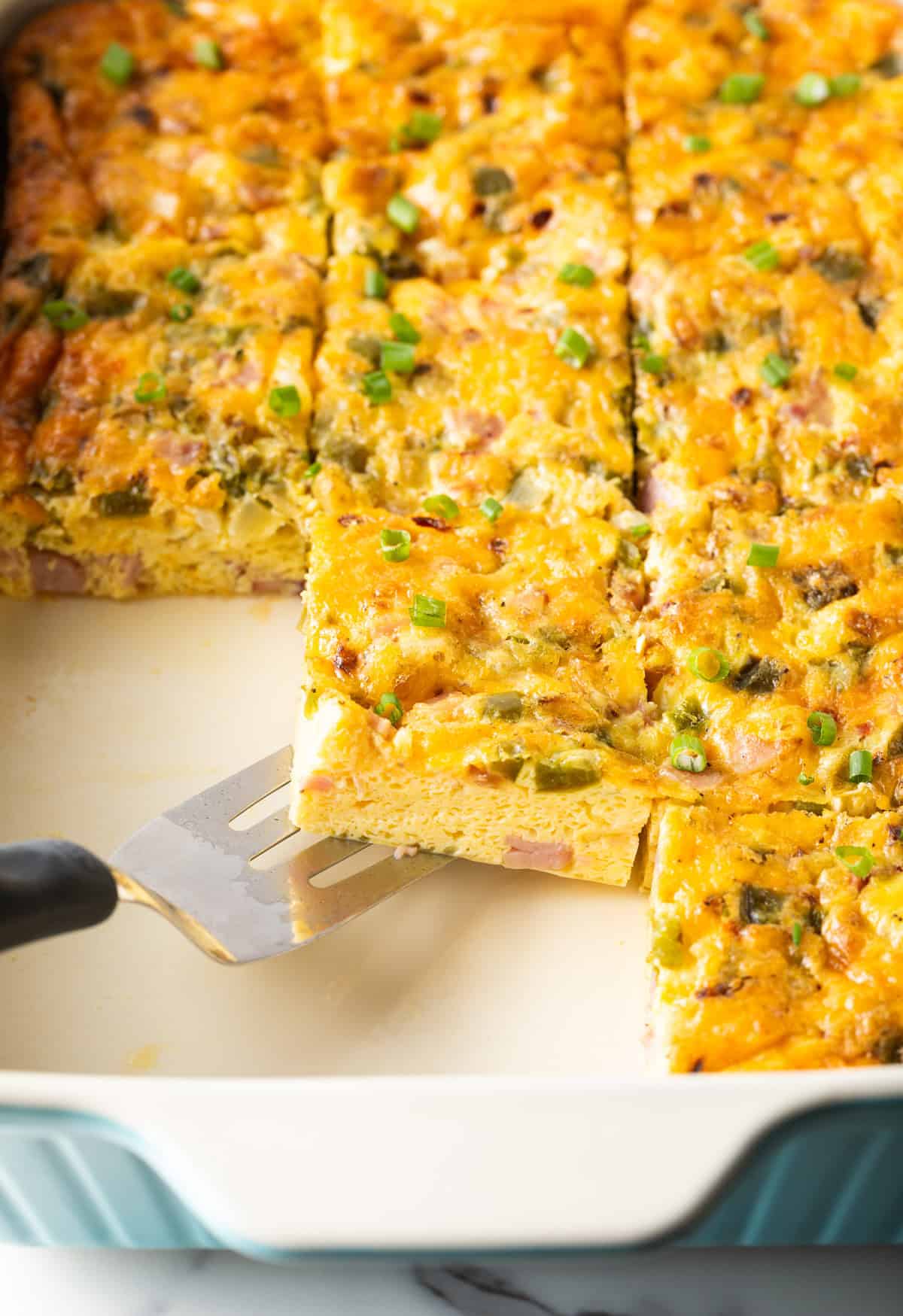 A slice of a western omelette casserole is being scooped by a metal spatula over a white baking tray filled with a Denver omelette casserole.