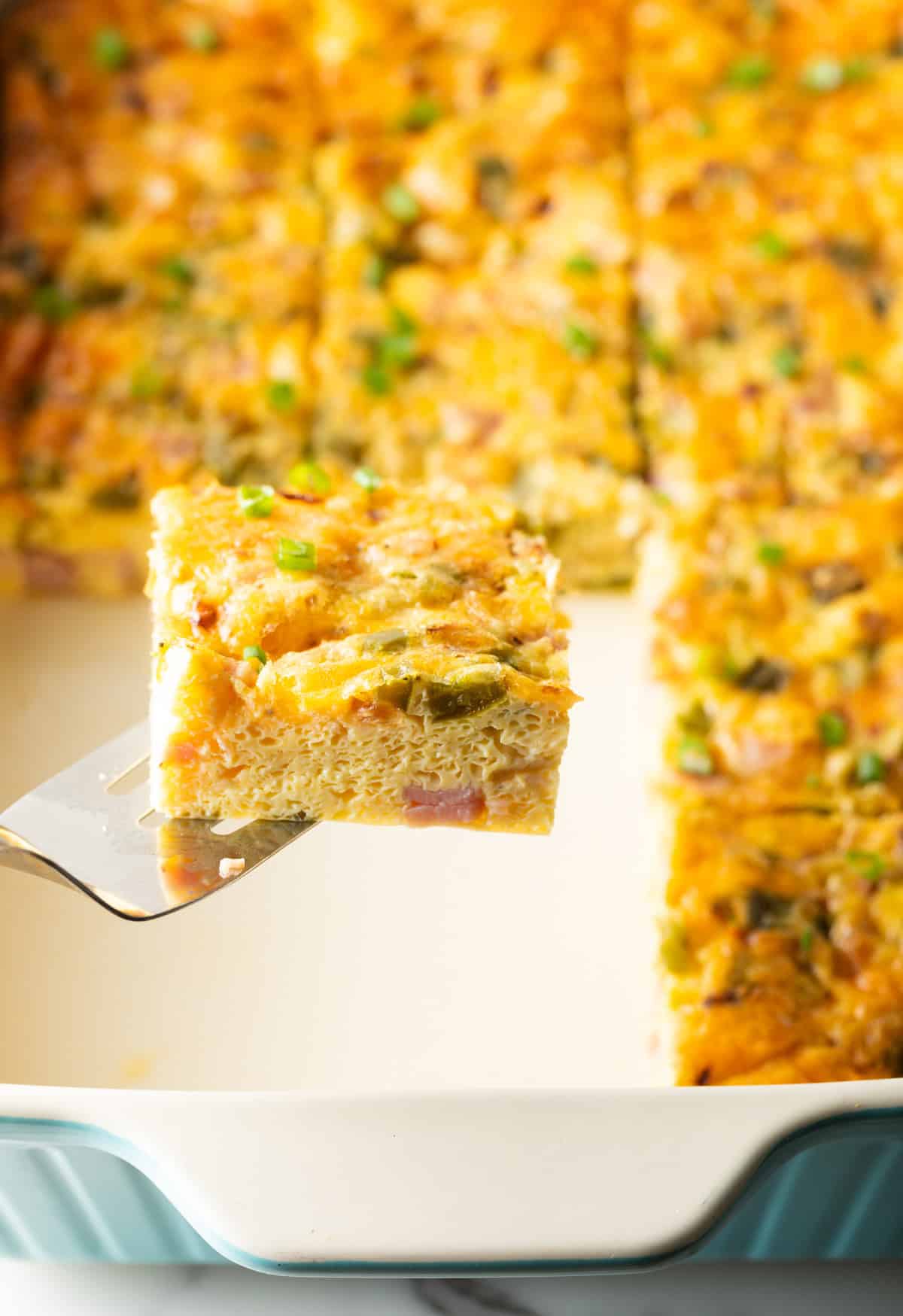 A slice of a Denver baked omelette is being held by a metal spatula over a white baking tray filled with a Denver omelet casserole.