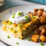 A slice of baked Denver omelet casserole topped with sour cream, next to a serving of home fries on a white plate.