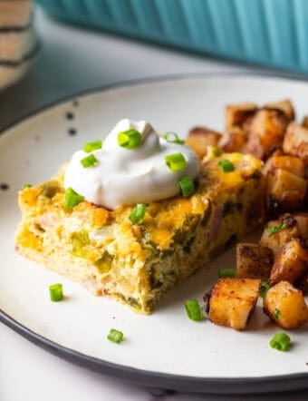 A slice of baked Denver omelet casserole topped with sour cream, next to a serving of home fries on a white plate.