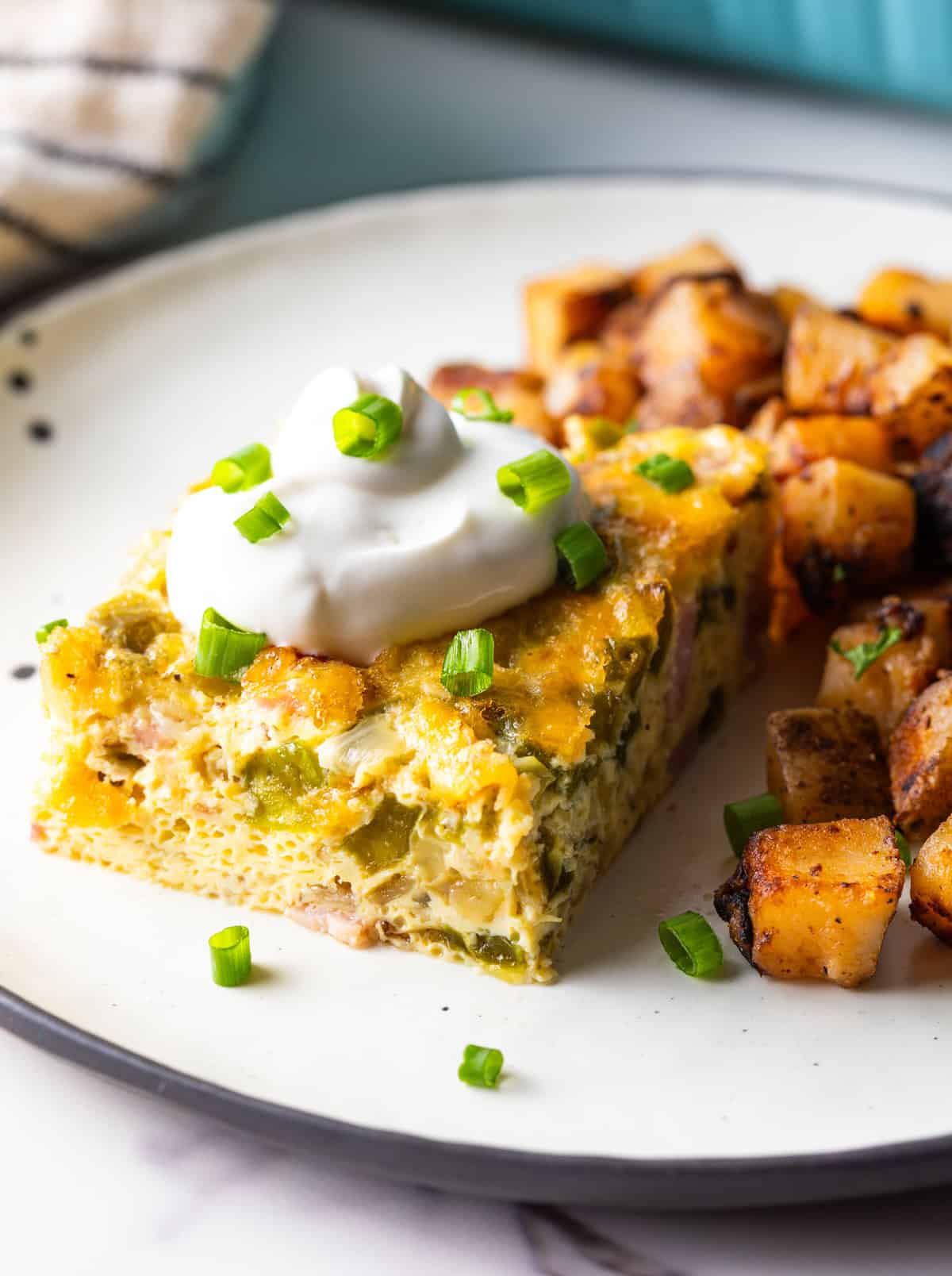 A slice of Denver baked omelet casserole topped with sour cream, next to a serving of home fries on a white plate.