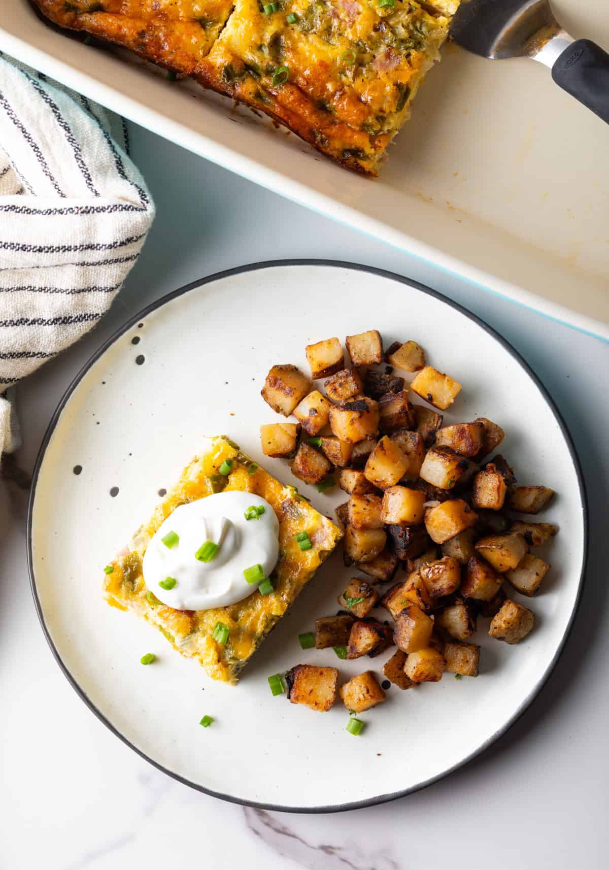 An overhead shot of a slice of baked Denver omelet casserole topped with sour cream, next to a serving of home fries on a white plate.