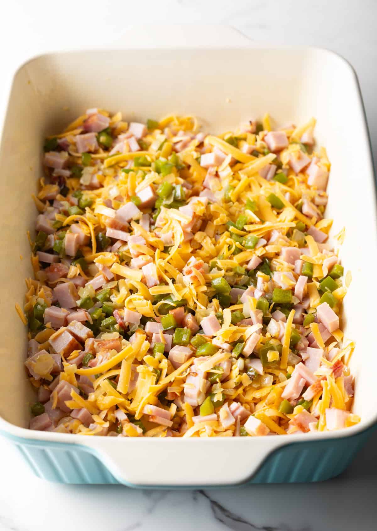 Shredded cheese is being mixed into a ham and vegetable mixture in a white baking dish.