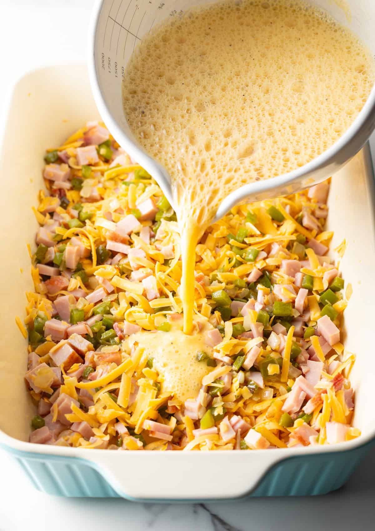 An egg mixture is being poured over ham, cheese, and vegetables, in a white baking dish.