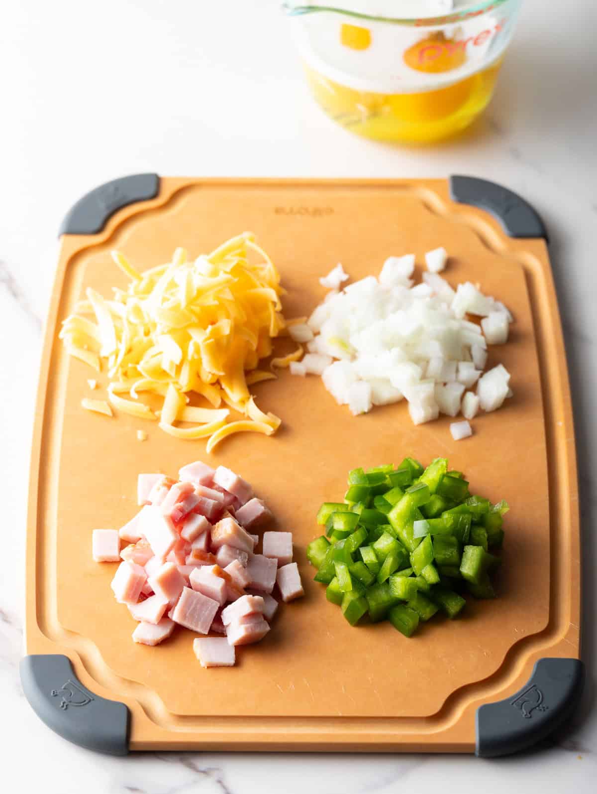 Diced onion, peppers, ham, and shredded cheese in four piles on a wooden cutting board.
