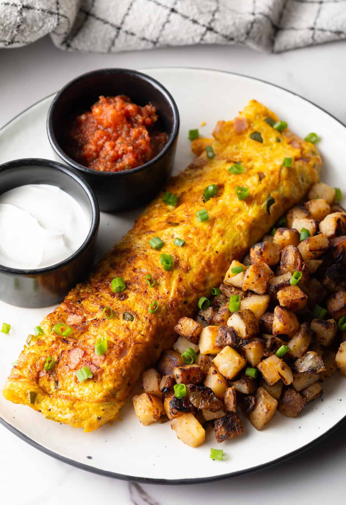 A Denver omelet between home fries and two black ramekins filled with sauce, on a white plate.