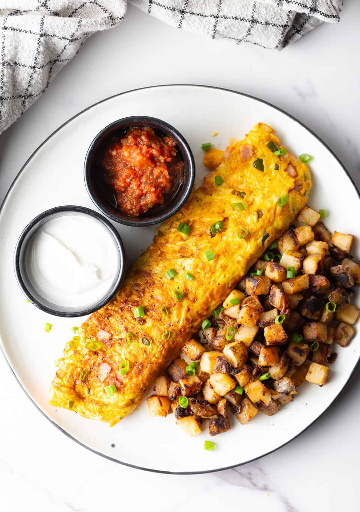An overhead view of a Denver omelet, breakfast potatoes, and ramekins of sauce, on a white plate.