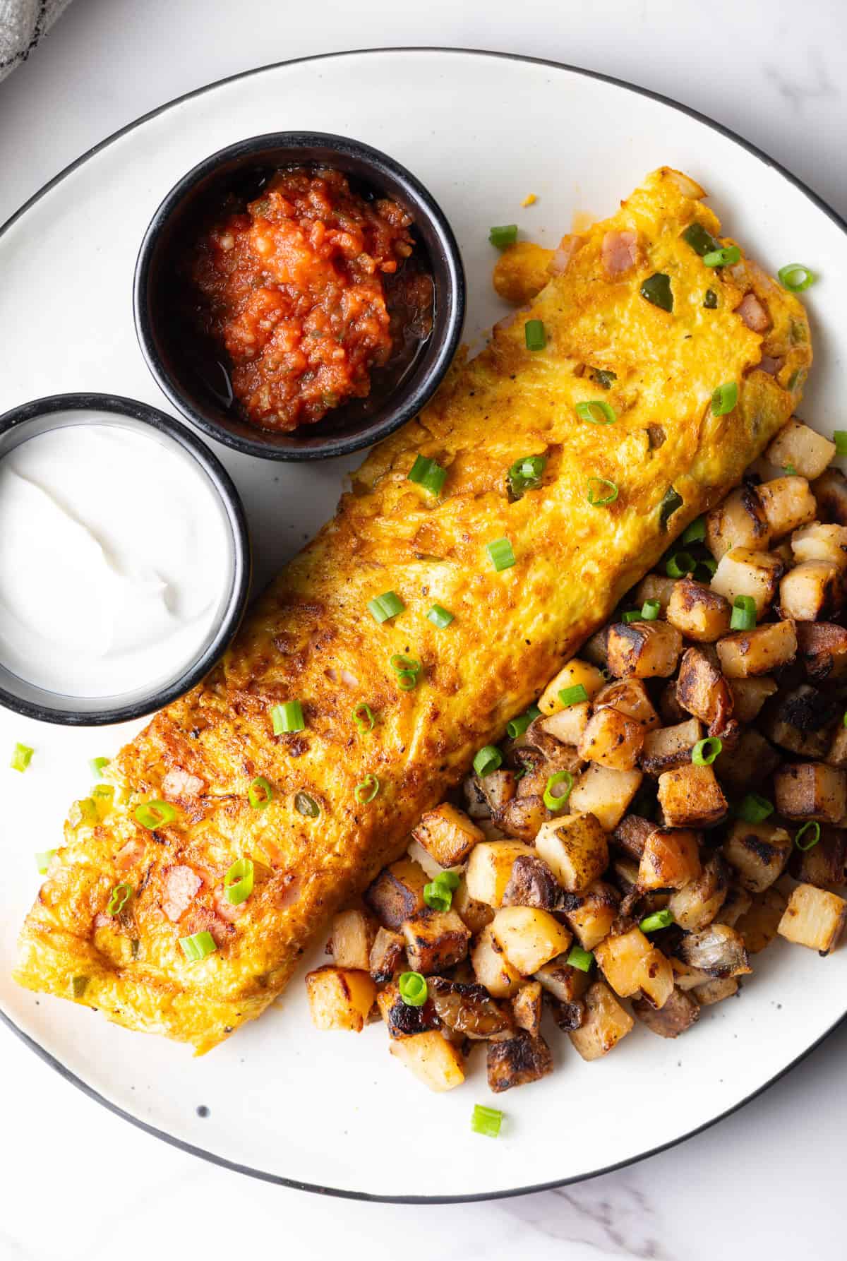 An overhead view of a Denver omelet, breakfast potatoes, and ramekins of sauce, on a white plate.