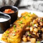 A Denver omelette between breakfast potatoes and two black ramekins filled with sauce, on a white plate.