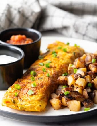 A Denver omelette between breakfast potatoes and two black ramekins filled with sauce, on a white plate.