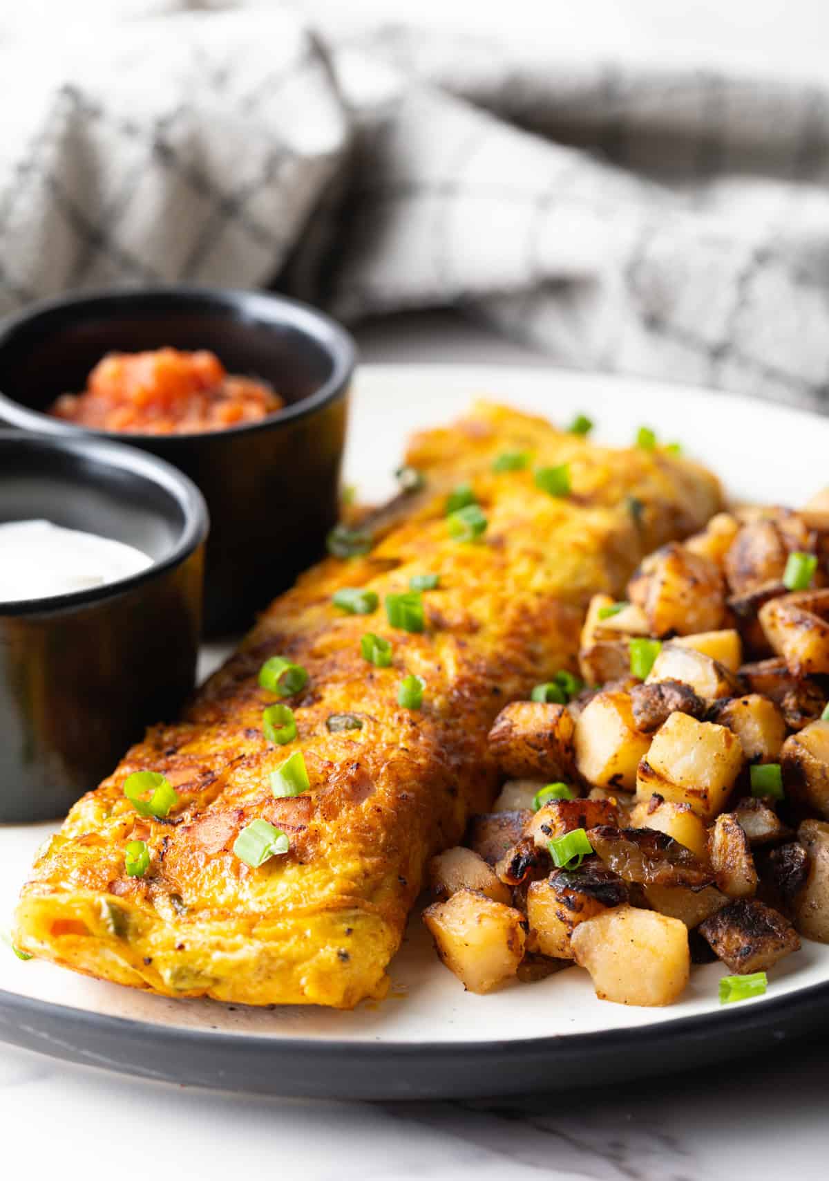 A Denver omelette between breakfast potatoes and two black ramekins filled with sauce, on a white plate.
