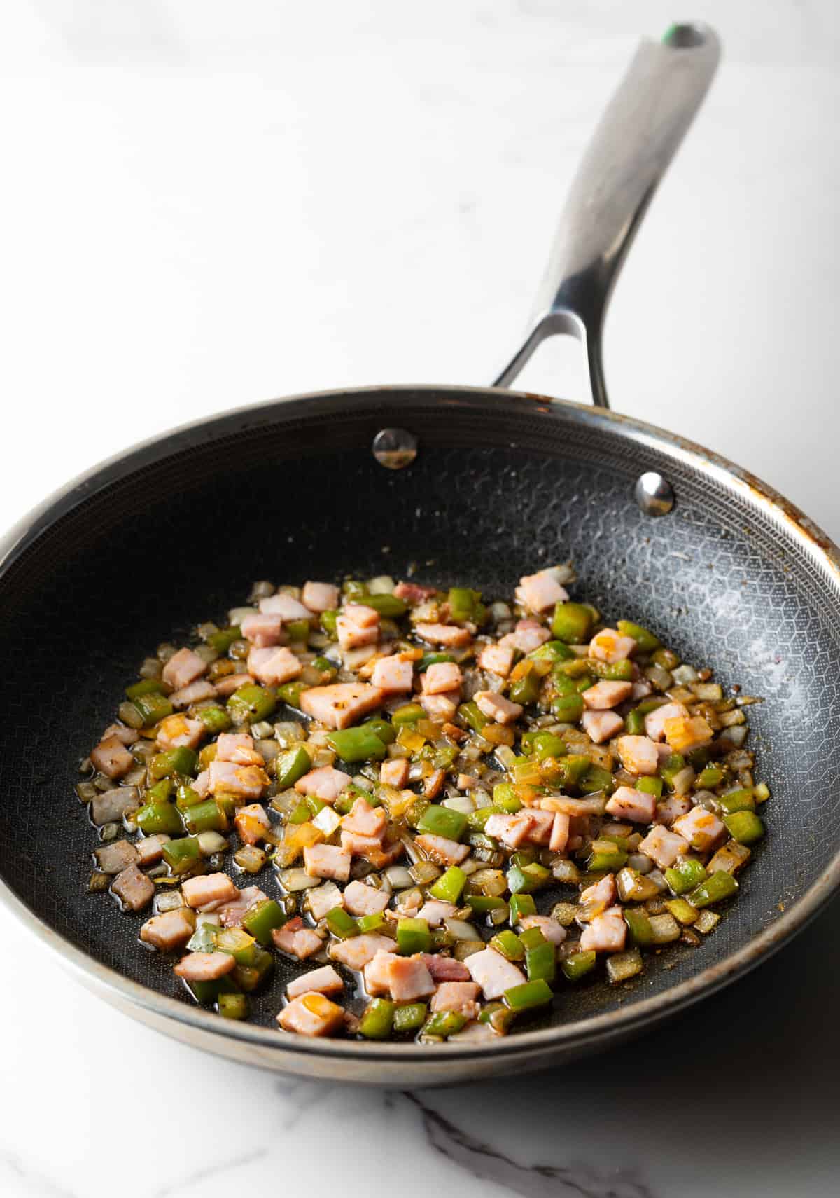 Diced onion, green peppers, and ham are being sautéed in a black pan.