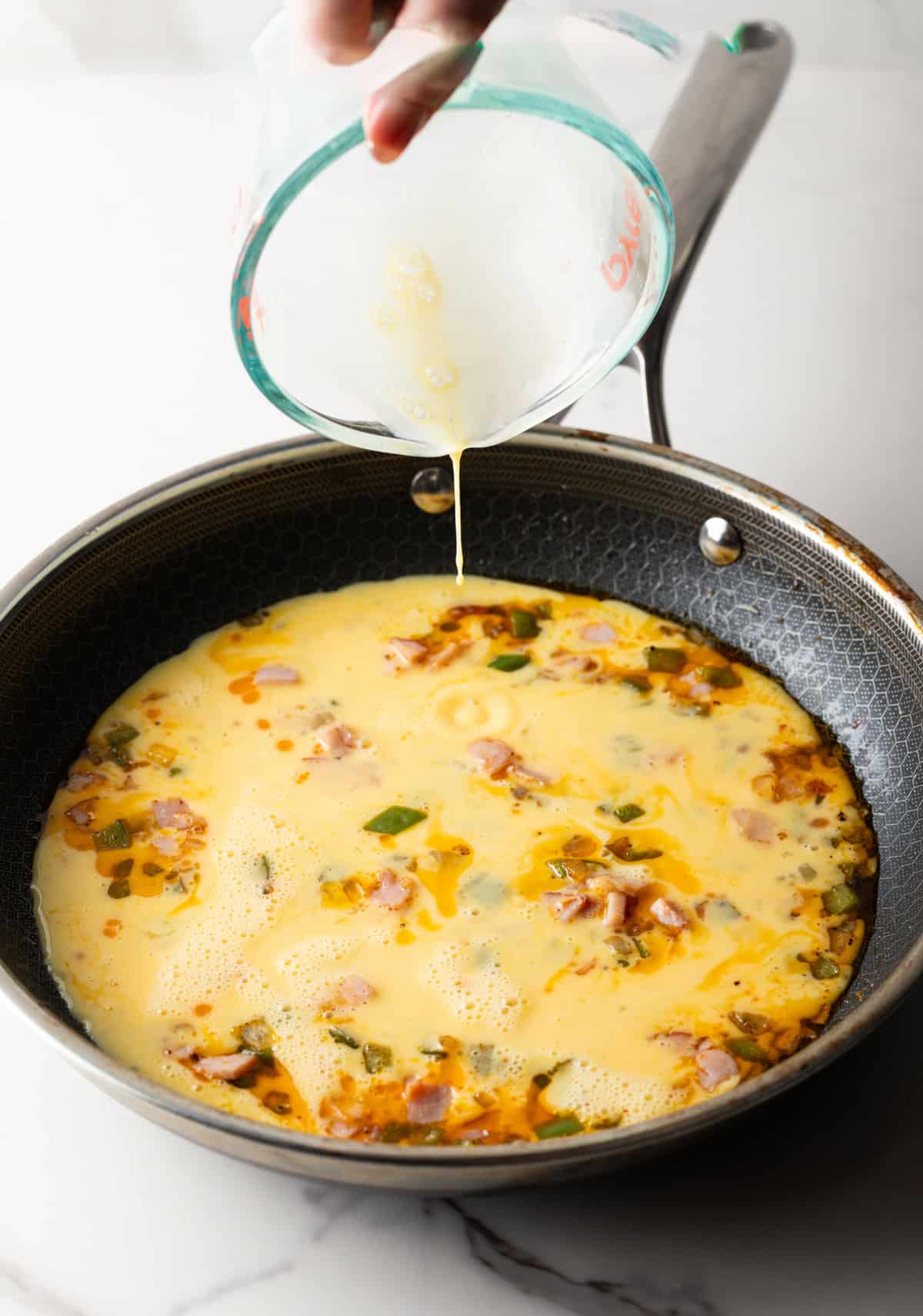 A cream and egg mixture is being poured into a black pan over sautéed vegetables and ham for a western omelet recipe.