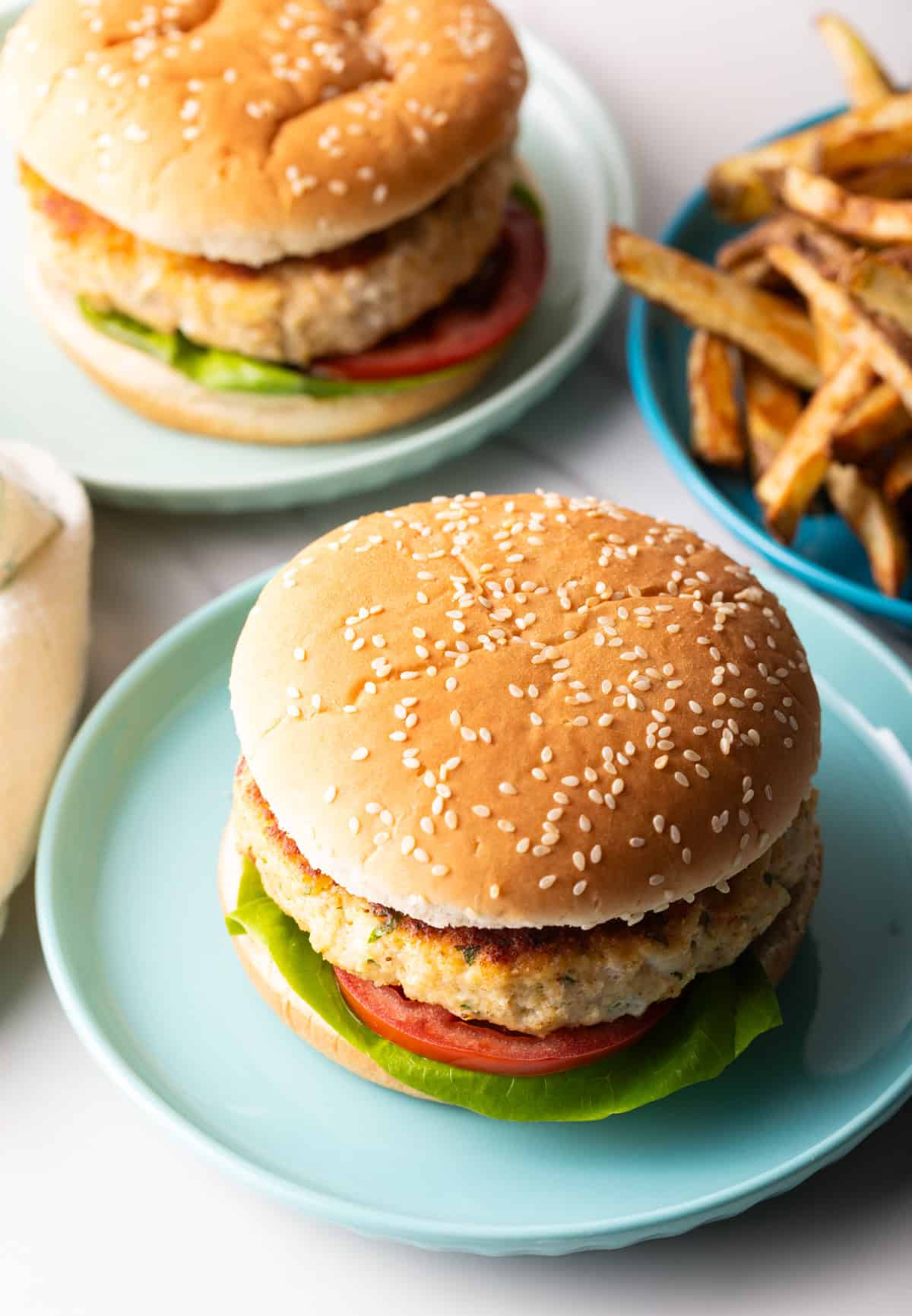 Homemade fish hamburgers with lettuce and tomato on blue plates, with a plate of fries on the side.