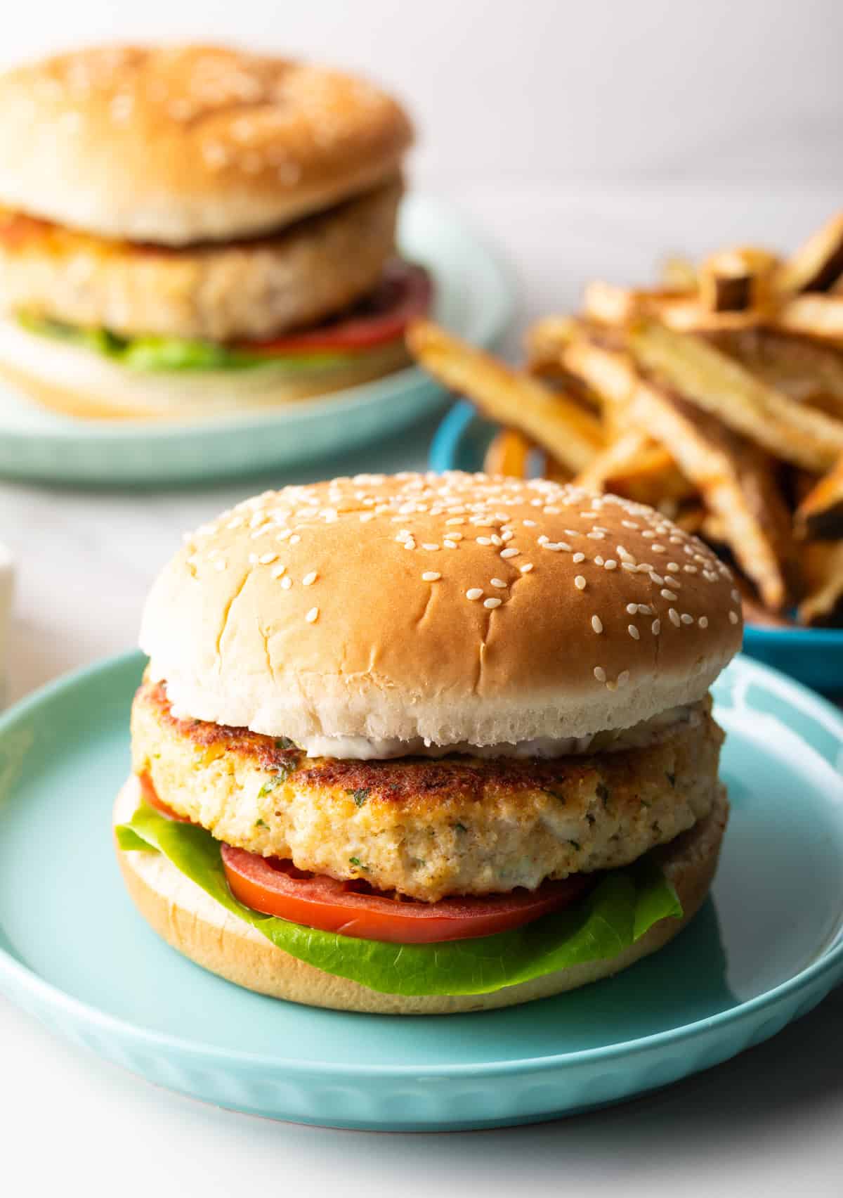 A homemade fish burger with lettuce and tomato on a blue plate, with a plate of fries on the side.