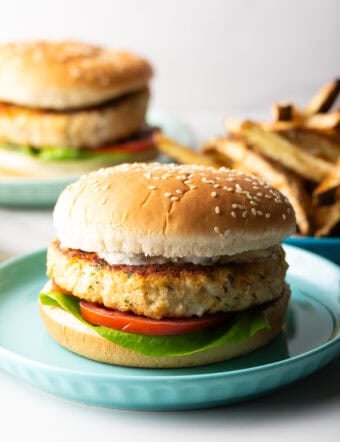 Homemade fish burgers with lettuce and tomato on blue plates, with a plate of fries on the side.