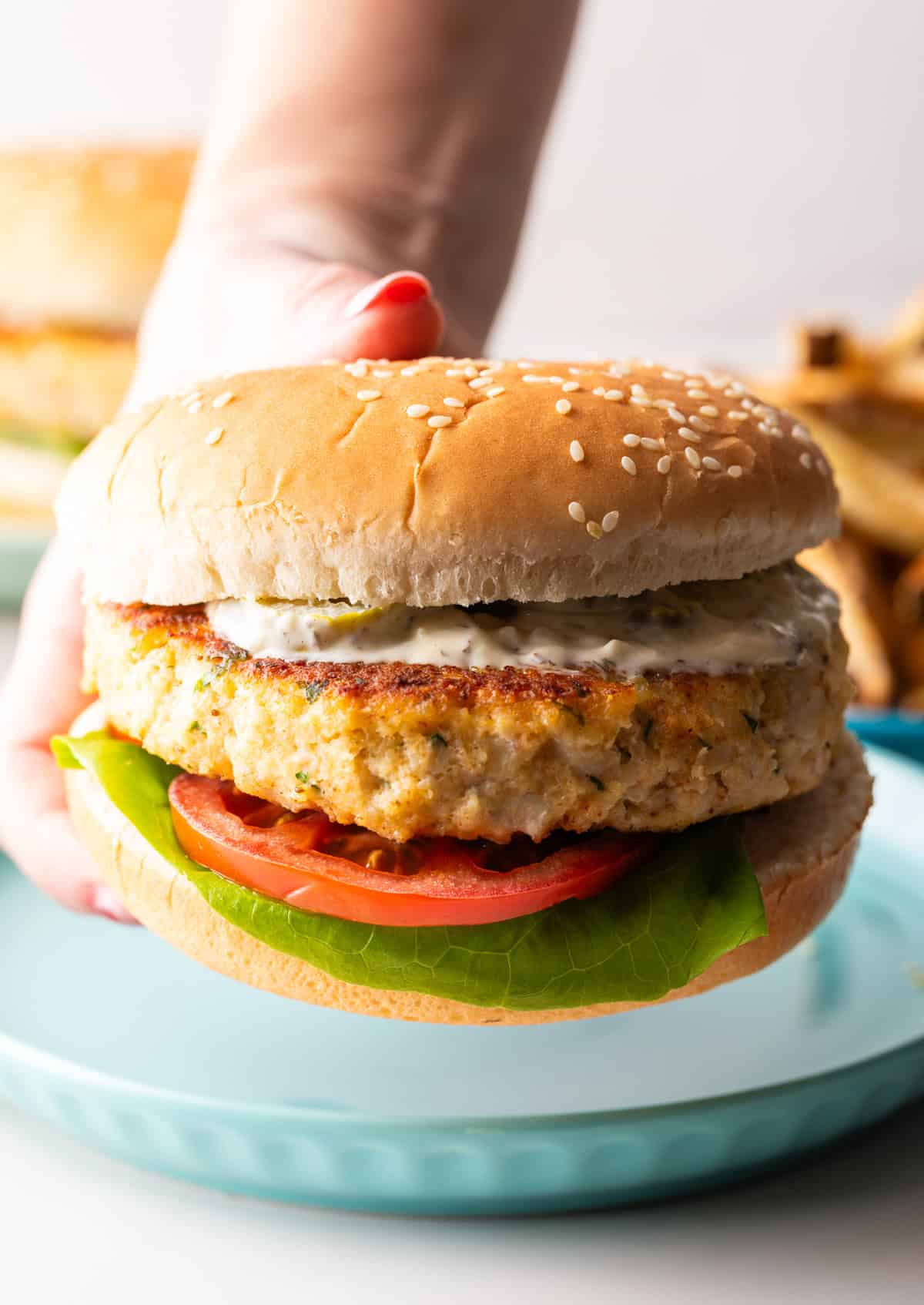 A hand is holding a fish hamburger above a blue plate.