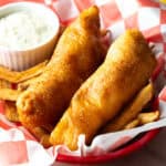 A crispy battered fried fish recipe in a paper lined basket, sitting on top of French fries.