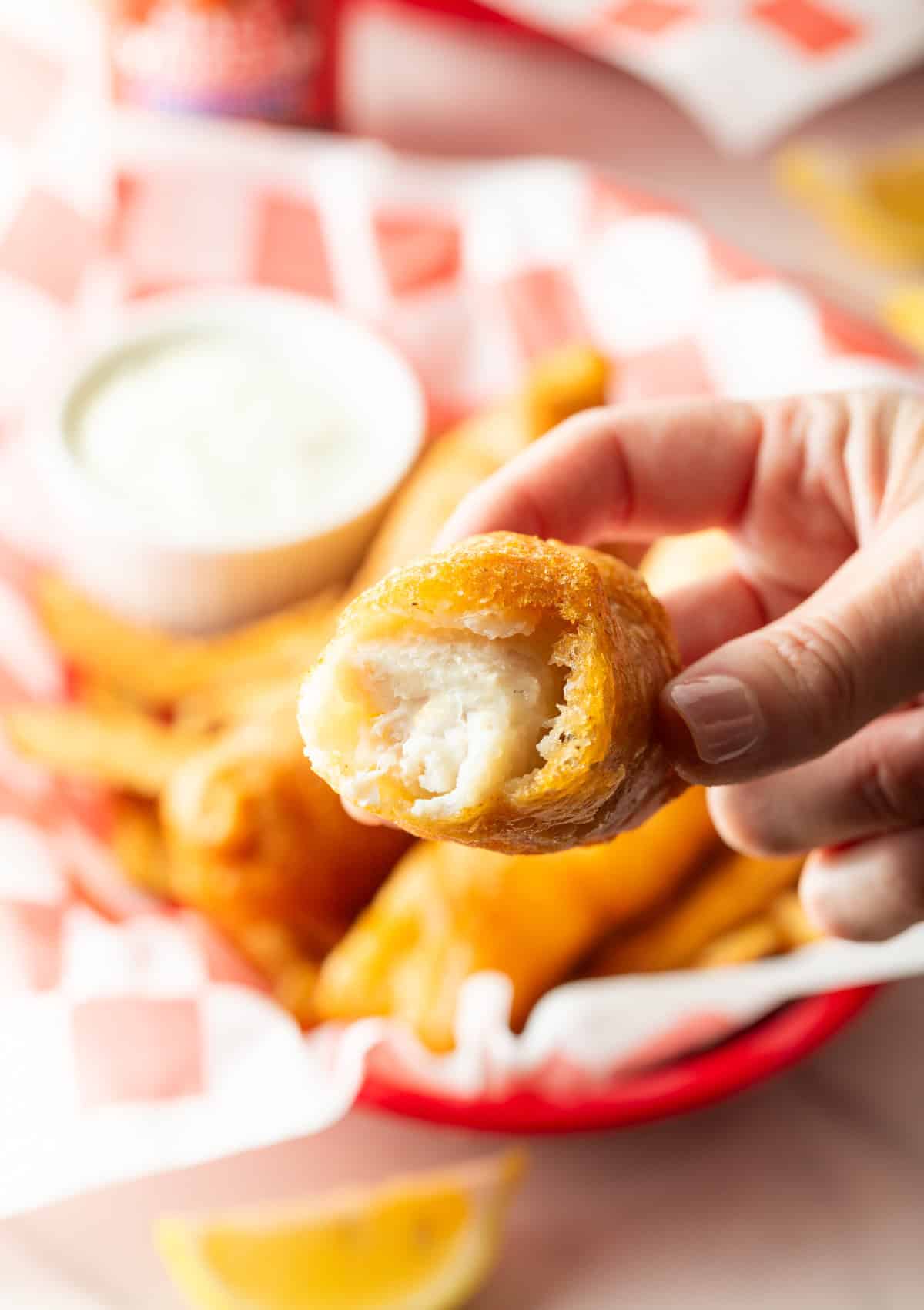 A hand is holding a piece of battered fried fish with a bite taken out, and it is being held over a basket of fish and chips.