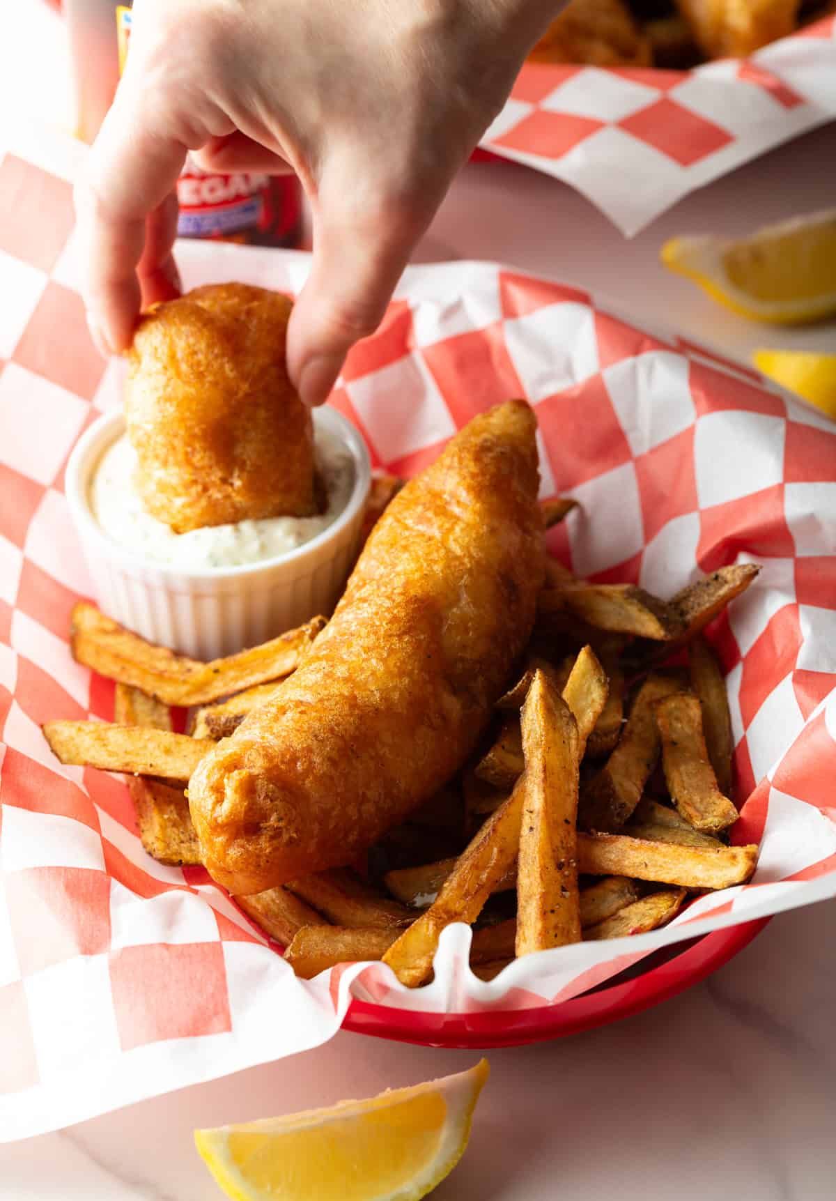 A hand is dunking a piece of crispy fried fish into a ramekin filled with sauce.