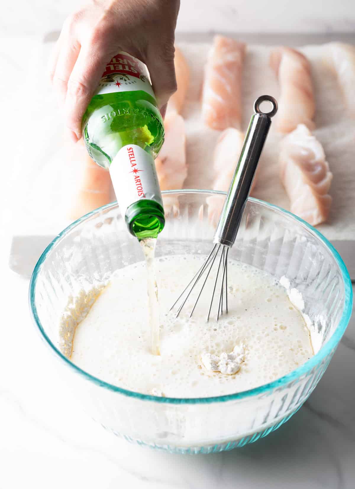 A bottle of beer being poured into a fried fish batter in a glass bowl.