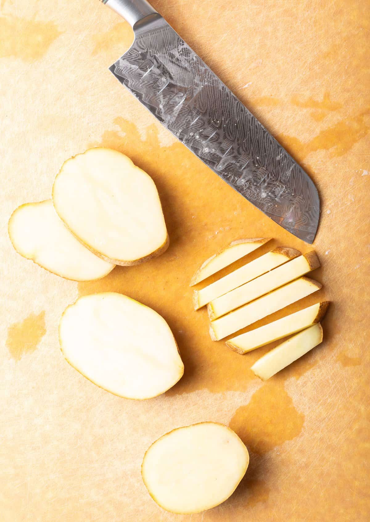 Top down view slices and sticks of raw potato on a cutting board with a large knife.