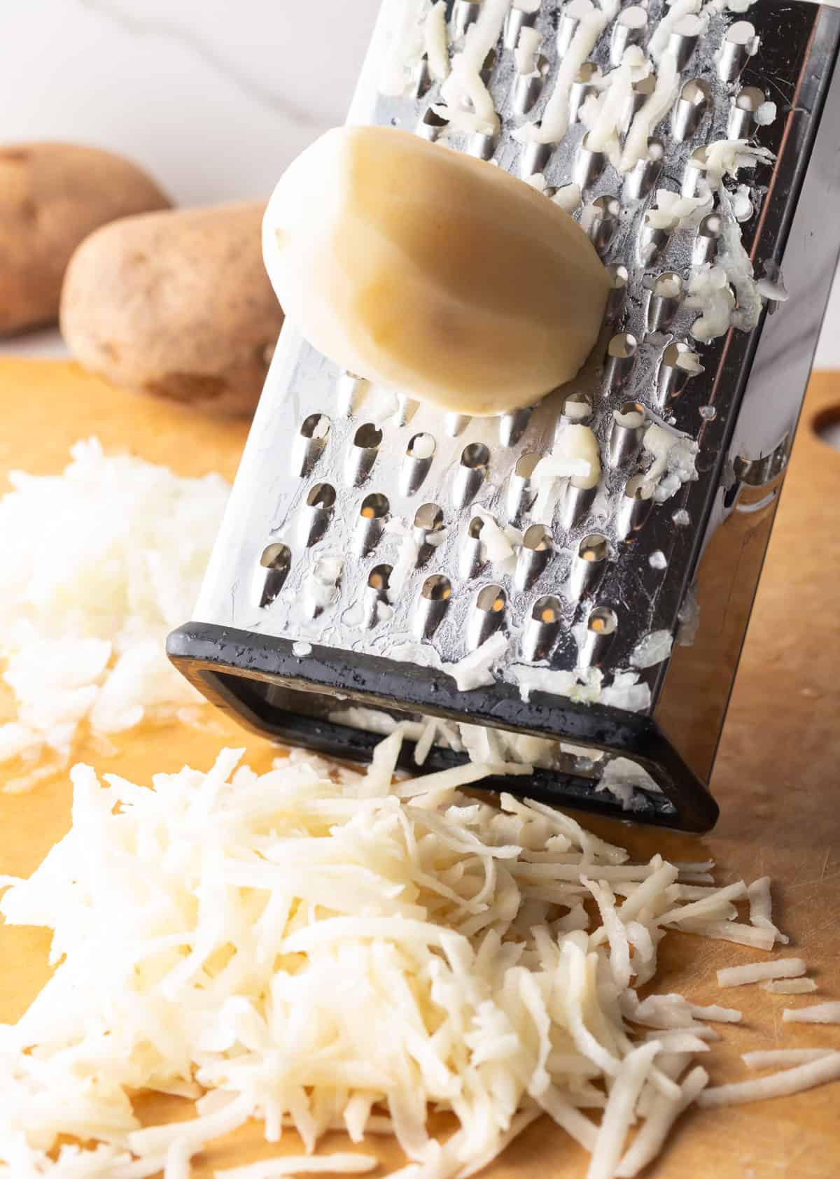 A peeled potato is being shredded with a box grater onto a wooden cutting board.