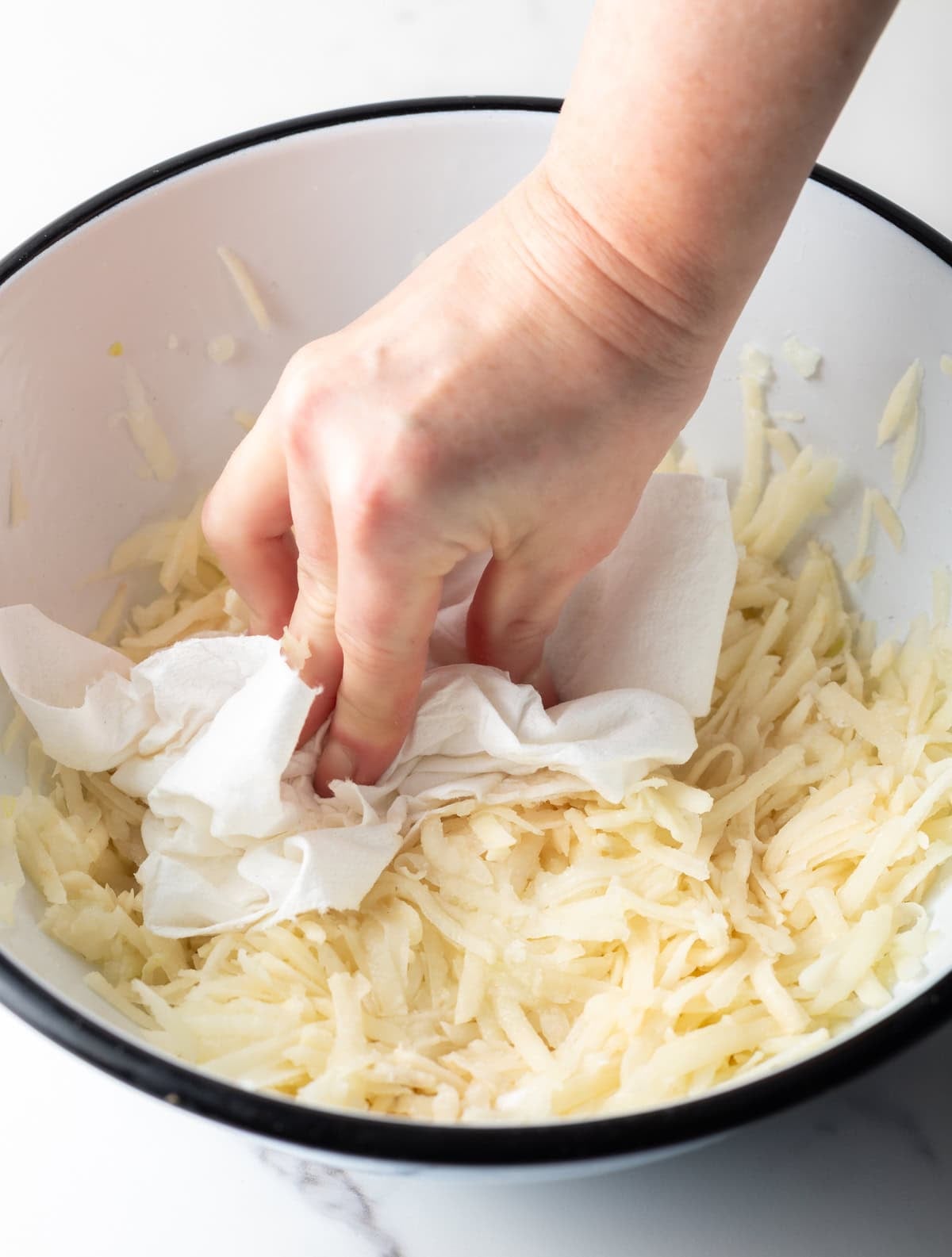 A hand holding a paper towel that is being used to remove moisture from shredded potatoes for a homemade hash brown recipe.