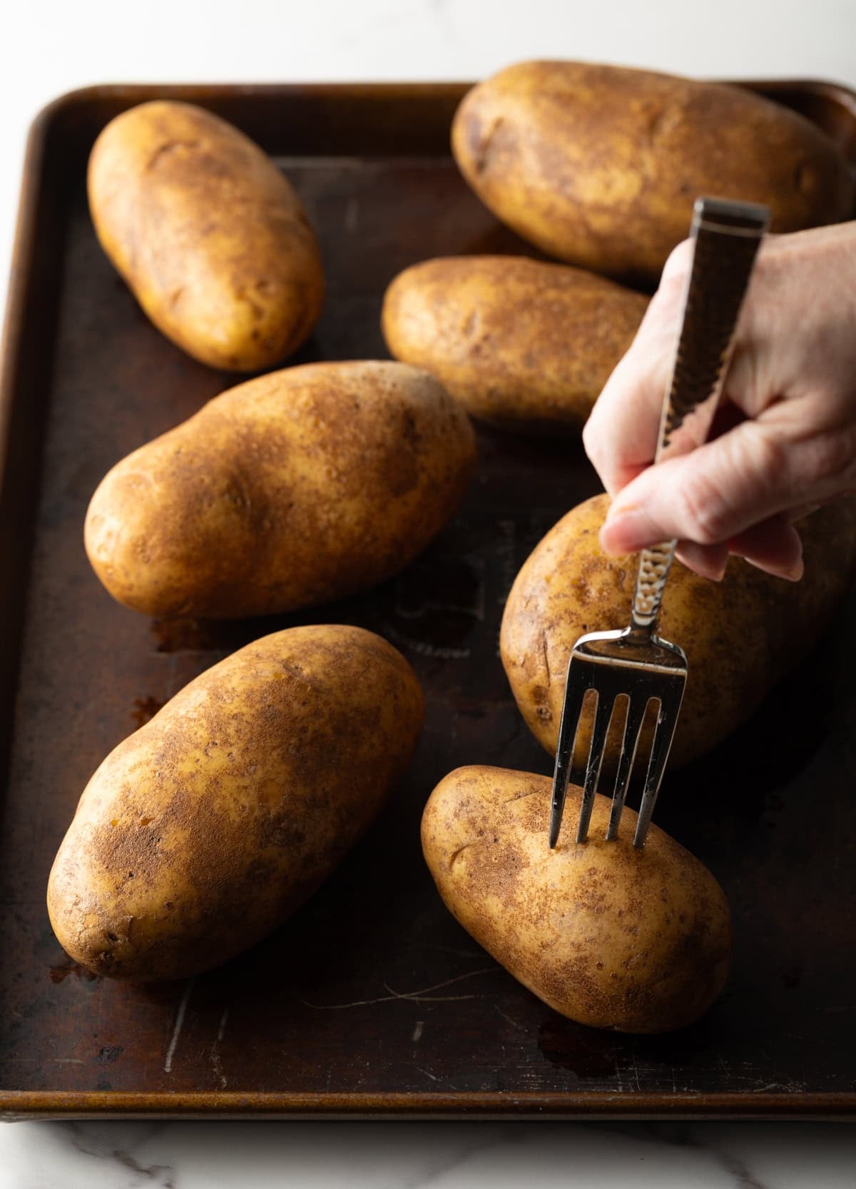 A fork is poking whole russet potatoes on a baking sheet.